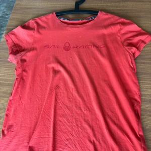 Röd t-shirt från Sail Racing - Röd t-shirt från Sail Racing med rund hals och tryckt logga på bröstet. Klassisk passform och korta ärmar, perfekt för en avslappnad stil.