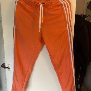 Orange träningsbyxor från Adidas - Säljer ett par orange träningsbyxor från Adidas med vita ränder längs sidorna och vit dragsko i midjan. Byxorna har en sportig look och är perfekta för gymmet eller löprundan. Klassisk Adidas-logga på ena benet. (Har en svart fläck på benet som visas i sista bilden)