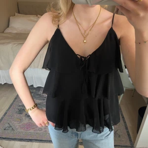 Svart volangig linnetopp - Säljer en svart linnetopp med tunna axelband och flera lager volanger. Toppen har en liten knytning framtill och är tillverkad i ett lätt, transparent material som ger en luftig känsla. Kom tyvärr aldrig till användning🖤