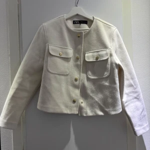Vit kavajjacka från Zara - Stilren vit kavajjacka från Zara med rund halsringning och guldknappar framtill. Jackan har två stora bröstfickor med lock och knappar. Klassisk rak passform och lång ärm. Perfekt för en trendig och clean look.