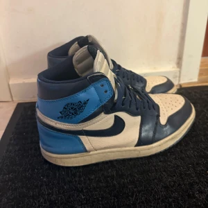Nike Air Jordan 1 High blå och vita sneakers - Säljer ett par Nike Air Jordan 1 High sneakers i blått, vitt och mörkblått skinn. Klassisk hög modell med snörning och perforerad tå. Ikonisk Air Jordan-logga på sidan och Nike-märke på plösen. Perfekta för dig som gillar streetwear.