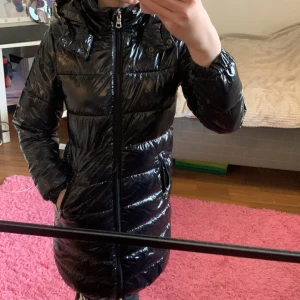 Svart glansig ljacka från Ellos💕 - Säljer en svart glansig lång pufferjacka från Ellos med huva och dragkedja framtill. Jackan har två fickor med dragkedja och quiltad design💗 går även att vika in så den blir kort💕