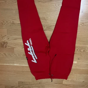 Röda mjukisbyxor med vit logga - Röda mjukisbyxor med vit broderad logga och text på vänster ben. Byxorna har snörning i midjan och är tillverkade i mjukt material. Perfekta för en avslappnad stil.