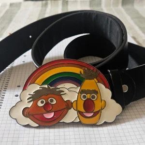 Svart bälte med färgglad spänne från Mupparna - Säljer ett svart bälte med ett spänne med motiv av två figurer från Mupparna. Bältet har en klassisk form och spännet är iögonfallande och lekfullt.