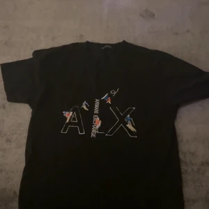 Svart t-shirt från Armani Exchange - Säljer en svart t-shirt från Armani Exchange med stort A|X-tryck och färgglada detaljer på bröstet. Klassisk passform och rund halsringning. Perfekt för dig som gillar stilrena märkesplagg.
