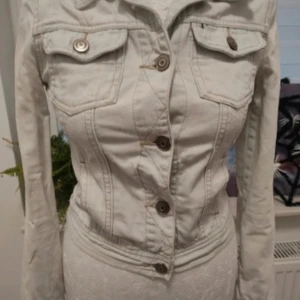 Vintage jeansjacka - Säljer en vintage vit jeansjacka med krage och två fickor framtill. Jackan har långa ärmar och stängs med stora metallknappar. Figursydd modell som ger en snygg siluett. Perfekt att slänga över en t-shirt eller klänning. (Detta var ai men allt hon sa var sant)