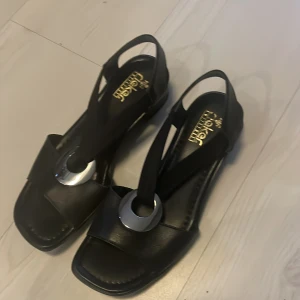 Svarta sandaler med metallring från Rieker - Snygga svarta sandaler från Rieker med bred rem över foten och en stor silverfärgad metallring som detalj framtill. Skorna har öppen tå och platt sula, perfekt för varma dagar. Klassisk och stilren design med bekväm passform.