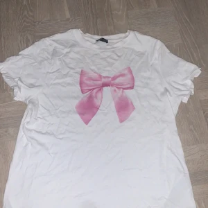 Vit t-shirt med rosa rosett från FB Sister - Vit t-shirt från FB Sister med en stor rosa rosett tryckt på bröstet. Modellen är regular fit och har korta ärmar. Perfekt för en söt och avslappnad stil.