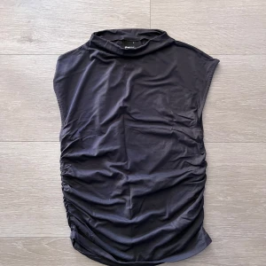 Mörkgrå ärmlös topp med rynkade sidor - Säljer en mörkgrå ärmlös topp med hög hals och rynkade sidor. Toppen har en stilren och modern look, perfekt att matcha med både jeans och kjol. Mjukt och stretchigt material för en bekväm passform.