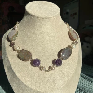 Halsband med lila stenar och pärlor - Unikt halsband med stora ovala stenar i lila och grå toner, varvat med runda lila stenar och vita pärlor. Silverfärgad infattning och kedja. Perfekt för dig som vill sticka ut med ett färgstarkt smycke. Smycket är flexibelt och går att forma efter sin egen hals.