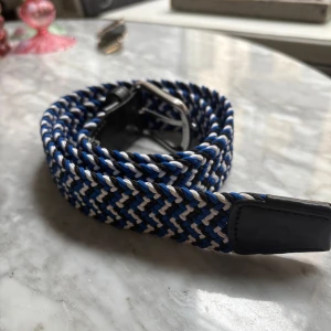 Flätat bälte från J.Lindeberg - Snyggt flätat bälte från J.Lindeberg i blått, vitt och svart med svarta skinn-detaljer och silverfärgat spänne. Bältet är 90 cm långt och passar midja 32-33 tum. Perfekt för att ge outfiten en sportig touch.