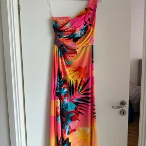 Färgglad långklänning från Lucas & Emma - Säljer en maxiklänning från Lucas & Emma med tropiskt blommönster i rosa, orange, blått och gult. Klänningen har en axelbandslös design och draperad överdel, perfekt för sommarens alla tillfällen.