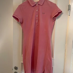 Rosa pikéklänning från Lacoste - Säljer en klassisk rosa pikéklänning från Lacoste med krage och knappar framtill. Klänningen har korta ärmar och är tillverkad i bomull. Perfekt för en avslappnad och sportig look.