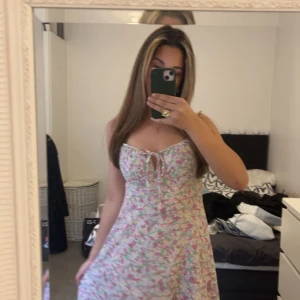 Blommig sommarklänning med knyt framtill - Söt blommig klänning med smala axelband och knyt framtill. Ljus botten med rosa och gröna blommor. Perfekt för den som gillar färgen 