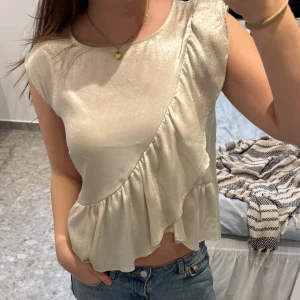 Champagnefärgad blus med volang - Champagnefärgad blus i glansigt material från DMK trend. Blusen har ett omlottliknande snitt framtill och volangdetalj längs nederkanten. Mycket bra skick!