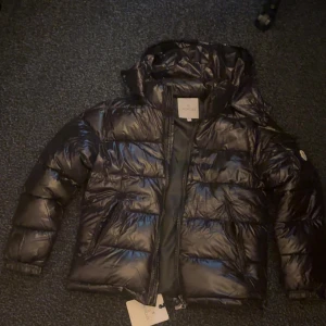 En fin moncler maya jacka  - Säljer en svart, glansig dunjacka från Moncler med huva och dragkedja. Helt oanvänd och nyskick. Jackan har klassisk pufferstil och Moncler-logga på ärmen. Perfekt för kyliga dagar och riktigt snygg till streetwear. 