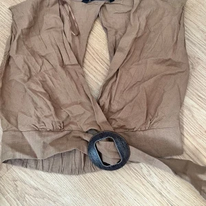 Beige omlottopp med bälte från Zara - Säljer en beige omlottopp från Zara i luftig bomull med bred resår i midjan och ett snyggt bälte med stor metallspänne. Toppen är ärmlös och har en djup v-ringning framtill. Perfekt för varma dagar!