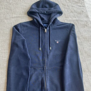 Marinblå hoodie från GANT med dragkedja - Skön GANT zip som är använd några gånger. Nypris ligger på 1500 så de är ett bra pris för kvaliteten. Den har små märken på armbågarna men det syns inte när man har på sig den. Storleken är XS, men den passar helt perfekt till S
