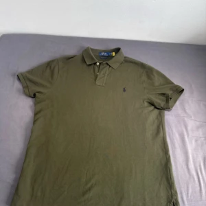 Olivgrön pikétröja från Ralph Lauren - Snygg olivgrön pikétröja från Polo Ralph Lauren i modellen Custom Slim Fit. Tröjan har klassisk krage, korta ärmar och två knappar framtill. Ikoniska lilla loggan broderad på bröstet. Perfekt för en clean och stilren look.