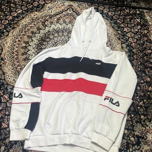Vit hoodie från FILA med ränder, strl L - Snygg vit hoodie från FILA i storlek L med breda svarta och röda ränder över bröstet och ärmarna. Tröjan har huva med snörning, känguruficka och tryckt FILA-logga på bröstet och ärmen. Perfekt för en sportig och avslappnad stil.