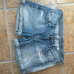 Blå jeansshorts med slitningar - Snygga blå jeansshorts med uppvikta ben och slitna detaljer framtill och baktill. Klassisk femficksmodell med knapp och dragkedja. Perfekta för varma dagar och ger en avslappnad vibe. Ljus tvätt och casual look.