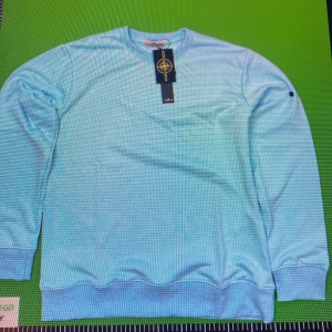 Ljusblå sweatshirt från Stone Island - Snygg ljusblå sweatshirt från Stone Island med klassisk rund hals och ribbade muddar. Tröjan har lång ärm och ikonisk logotyp-patch vid halsen. Perfekt för dig som gillar stilrena och trendiga plagg med streetkänsla.