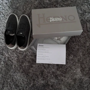 Svarta sneakers från Herno i mocka - Snygga svarta sneakers från Herno i mjuk mocka med vita sulor och bruna detaljer med logomönster på hälen. Klassisk rund tå, snörning och stilren design som passar perfekt till jeans eller chinos.