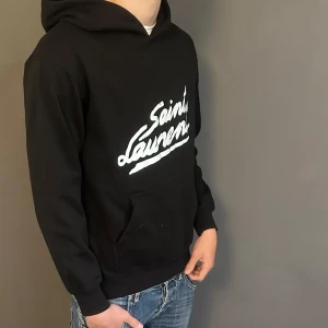 Svart Saint Laurent hoodie med tryck - Svart hoodie från Saint Laurent med vit logga framtill. Klassisk känguruficka, huva och ribbade muddar. Mjuk insida och avslappnad passform, perfekt för en cool streetstyle-look.