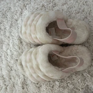 Fluffiga UGG sandaler i vitt och rosa - Supermysiga fluffiga sandaler från UGG i vitt med ljusrosa detaljer och breda remmar. Sulan är i gummi med UGG-logga undertill och ett elastiskt band bak för extra support. Perfekta för att chilla hemma eller när du vill ha något riktigt mjukt på fötterna.
