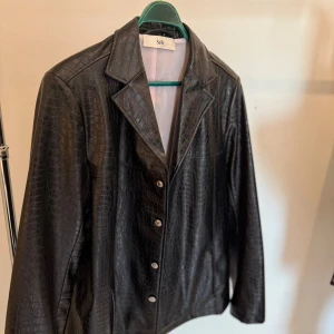 Svart skinnjacka Séfr croco XL - Säljer en svart skinnjacka från Séfr i storlek XL med croco-mönster och klassisk krage. Jackan har knappar framtill och är tillverkad i syntetmaterial med en glansig finish. Perfekt för dig som vill sticka ut med en edgy vibe.