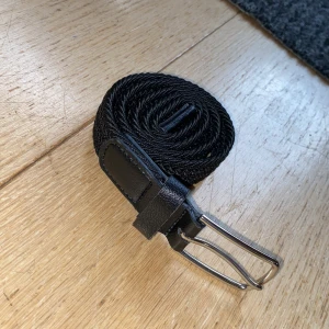 Svart flätat bälte med skinn och metallspänne - Snyggt svart bälte med flätad design och detaljer i svart skinn vid spännet. Spännet är i blank metall och bältet är flexibelt tack vare den flätade strukturen. Perfekt för att ge outfiten en stilren touch.