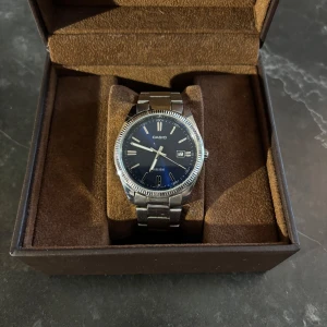 Casio klocka med blå urtavla - Stilren Casio klocka med blå urtavla och datumvisning. Klockan har en rund boett i silverfärgad metall och matchande länkarmband i rostfritt stål. Klassisk design med tydliga index och vridbar krona på sidan. Perfekt accessoar för dig som gillar enkel och snygg stil. 