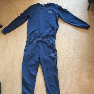 Blå EA7 dress med logga - Snygg blå dress från EA7 Emporio Armani med vit logga på benet och bröstet och svarta detaljer längs sidorna. Byxorna har resår och snörning i midjan samt muddar vid bensluten. Ingen huva på tröjan.