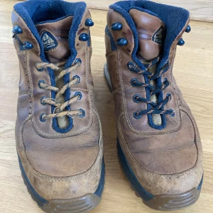 Nike ACG bruna kängor strl 43 - Nike ACG kängor i brunt skinn med blå detaljer och grov sula. Klassisk outdoor-vibe med snörning och robust känsla, perfekta för äventyr eller streetstyle. Märke och logga syns tydligt på plösen. Snygg kontrast mellan brunt och blått.