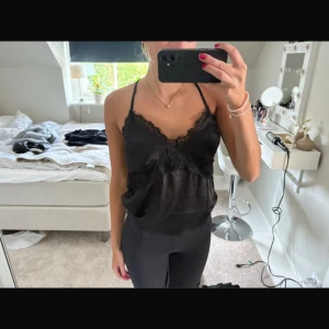 Svart linnetopp med spetsdetaljer - Snygg svart linnetopp med tunna korsade axelband och spets längs v-ringningen. Toppen har en lätt glansig finish och sitter löst över kroppen. Perfekt att styla med jeans eller kjol för en trendig look.