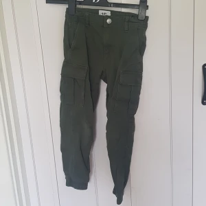 Gröna cargobyxor från 157, stl 130 - Mörkgröna cargobyxor från 157 med elastiska muddar vid bensluten och flera stora fickor på sidorna. Byxorna har normal passform och är tillverkade i mjuk bomull, perfekta för en avslappnad och cool stil.