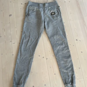 Grå mjukisbyxor från Philipp Plein, strl 14-15 - Grå mjukisbyxor från Philipp Plein med elastisk midja och muddar vid benslut. Byxorna har två fickor fram, en metallbadge med logga och ett stort tryckt P på baksidan. Strl 14-15, passar 150-170 cl eller xs