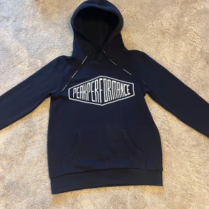 Mörkblå hoodie Peak Performance XS - Snygg mörkblå hoodie från Peak Performance i storlek XS. Stor vit logga framtill, huva med snörning och klassisk känguruficka. Perfekt för dig som gillar en sportig och avslappnad stil.