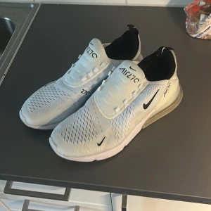 Nike Air Max 270 vita sneakers - Nike Air Max 270 sneakers i vitt med svarta detaljer och stor synlig Air-sula i hälen. Ovandelen är i ventilerande mesh och syntet, med klassisk Nike-logga på sidan och AIR270-tryck på plösen. Perfekta för dig som gillar sportig och modern stil. Dom är använda ett fåtal gånger.