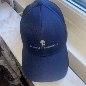 Blå keps från Brunello Cucinelli - Snygg marinblå keps från Brunello Cucinelli med broderad logotyp framtill. Klassisk böjd skärm och justerbar passform. Tillverkad i ett slitstarkt bomullsmaterial som ger en exklusiv känsla. Perfekt accessoar för en clean och stilren look.