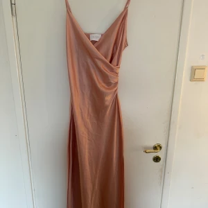 Champagnefärgad långklänning Sisters Point - Snygg långklänning från Sisters Point i en champagnefärgad nyans. Klänningen har smala axelband, omlott framtill och en snygg slits på sidan, ett glansigt, satinliknande material som ger en lyxig känsla. Perfekt som balklänning eller liknande.