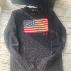 Mörkblå stickad tröja Polo Ralph Lauren - Mörkblå stickad tröja från Polo Ralph Lauren med amerikansk flagga i rött, vitt och blått på bröstet samt RL-broderi. Klassisk rund halsringning och långa ärmar. Perfekt för dig som gillar preppy stil och vill sticka ut lite extra.