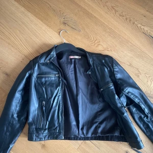 Svart skinnjacka från Saint Tropez - Snygg svart skinnjacka från Saint Tropez med flera fickor. Jackan har klassisk bikerstil med krage och lång ärm. Passar till allt och ger outfiten en grym attityd.