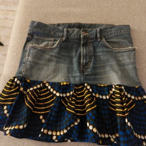 Jeanskjol med färgglad volang - Unik kort jeanskjol med volang i blått, gult och vitt mönster. Överdelen är klassisk denim med fickor och knapp, medan nederdelen har ett färgstarkt tyg med grafiskt mönster. Perfekt för dig som vill sticka ut med din stil.