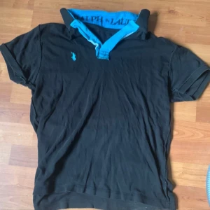 Svart pikétröja från Ralph Lauren - Svart pikétröja från Ralph Lauren med klassisk turkos krage och matchande logga på bröstet. Tröjan har korta ärmar och tydlig märkning på insidan av kragen. Perfekt för dig som gillar stilren och sportig look.