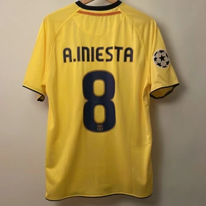 FC Barcelona Iniesta Champions League tröja - Gul FC Barcelona fotbollströja replika från Nike med A. Iniesta och nummer 8 på ryggen. Champions League-märke på ärmen, klubbmärke och Unicef-tryck på bröstet samt röd och mörkblå rand. Tillverkad i lätt och ventilerande polyester. Endast handtvätt gäller