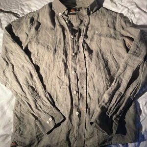 Grå linneskjorta från Morris M - Snygg grå skjorta i 100% linne från Morris med klassisk button down-krage och vita knappar. Broderad logga på bröstet ger en clean look. Perfekt för dig som gillar stilrena och luftiga plagg till sommaren. Dessvärre har den blekningar i armhålorna (bild 4/5). 