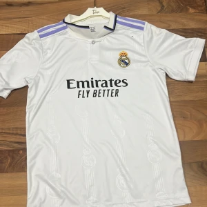 Real Madrid Vini Jr. 20 matchtröja - Real Madrid hemmatröja med VINI JR. och nummer 20 på ryggen. Vit med lila Adidas-ränder på axlarna, broderat klubbmärke och svart Emirates-tryck på bröstet. Tillverkad i lätt och ventilerande polyester med subtila klubbemblem i tyget.