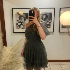 Mörkgrå one shoulder volangklänning från by malina - By malina klänning i modellen constance i grå st S och endast använd en gång eftersom jag hade den på min bal och därav är den i nyskick, köpt för 3500 men säljer för 1500❤️ klänningen är mörkgrå kortklänning med volanger och en axel. Klänningen har ett lager av transparent tyg, ballongärm på ena sidan och omlott-effekt över bysten. Perfekt för dig som vill sticka ut med en unik och trendig look.