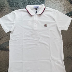 Vit Moncler piké med rödblå krage - Snygg vit pikétröja från Moncler med klassisk krage och knäppning framtill. Kragen har stilrena rödblå ränder och bröstet pryds av Monclers ikoniska logga. Tillverkad i bomull för en fräsch och bekväm känsla. Perfekt för dig som gillar stilren och sportig look.